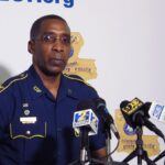 A Louisiana police officer speaking at a press conference with multiple cameras focused on him.