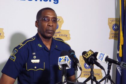 A Louisiana police officer speaking at a press conference with multiple cameras focused on him.