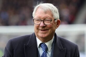 Alex Ferguson on a football field, happy and smiling brightly while enjoying the moment.