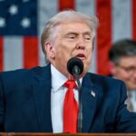 Donald Trump speaking into a microphone while wearing an elegant suit with the United States flag behind him.