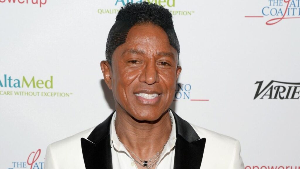 Jermaine Jackson smiling on a red carpet with a big smile and an elegant suit.