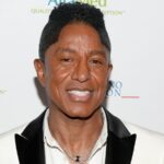 Jermaine Jackson smiling on a red carpet with a big smile and an elegant suit.