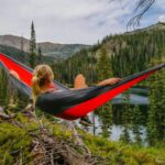 A woman sitting in a hammock tied between two trees while overlooking a landscape filled with nature.