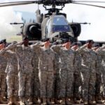 Many American soldiers saluting with their hands on their foreheads, with several military helicopters behind them.
