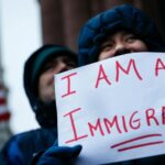 A person with their face covered holding a sign that says “I’m an immigrant.”