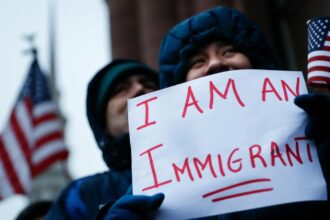 A person with their face covered holding a sign that says “I’m an immigrant.”