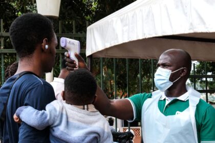 A man taking another person’s temperature while the person holds his child in his arms.