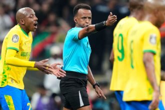 The Somali referee blowing the whistle for a foul against a team in yellow, whose players are protesting his decision