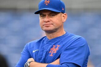 The now-former Mets manager sitting in his team's dugout, watching the game with a disappointed expression