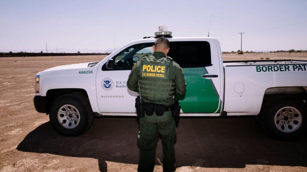 A U.S. Border Patrol officer standing beside his white and green patrol vehicle.