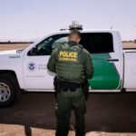 A U.S. Border Patrol officer standing beside his white and green patrol vehicle.