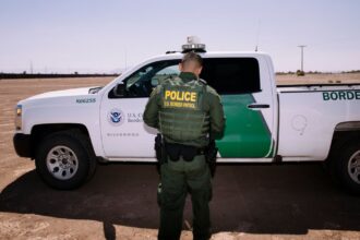 A U.S. Border Patrol officer standing beside his white and green patrol vehicle.