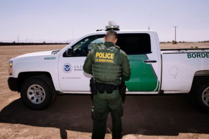 A U.S. Border Patrol officer standing beside his white and green patrol vehicle.