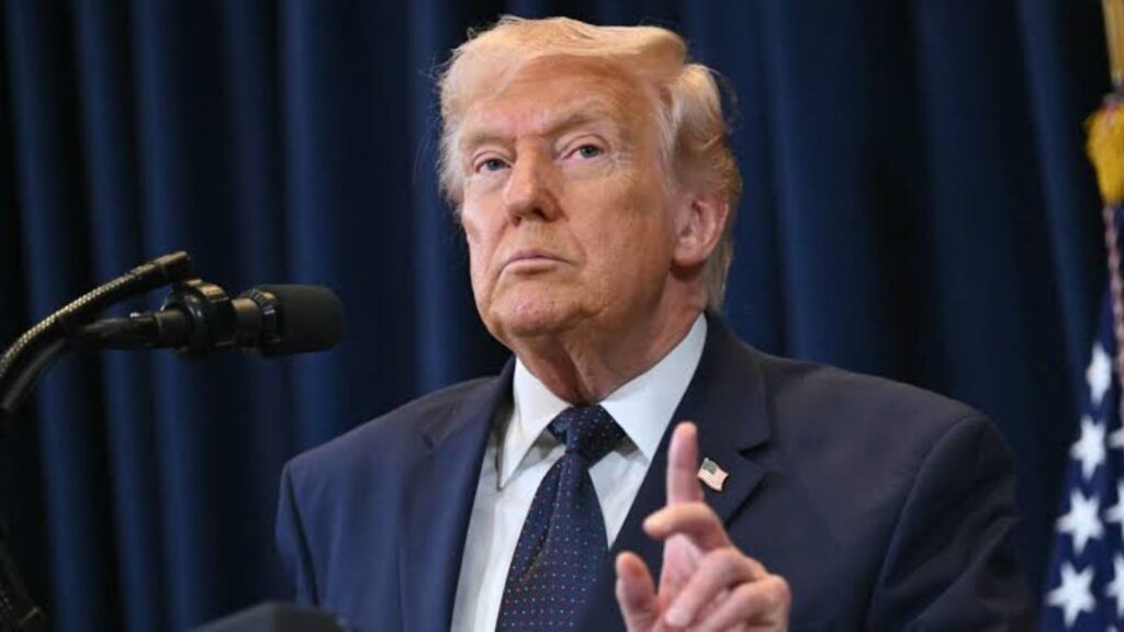 Trump standing in front of cameras and microphones, about to speak while wearing an elegant suit