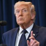 Trump standing in front of cameras and microphones, about to speak while wearing an elegant suit