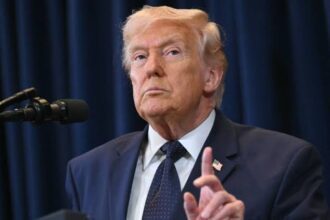 Trump standing in front of cameras and microphones, about to speak while wearing an elegant suit