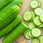 Several cucumbers stacked on a table, with one cucumber sliced into pieces beside them and a knife placed nearby