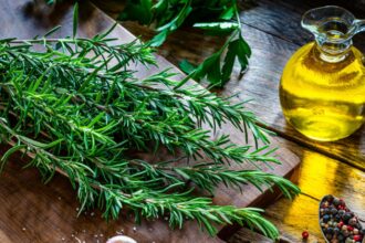 A large sprig of rosemary placed on a wooden kitchen cutting board, with a bottle of rosemary oil beside the sprig