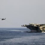 A massive U.S. aircraft carrier sailing across the sea while a helicopter closely follows it from the air