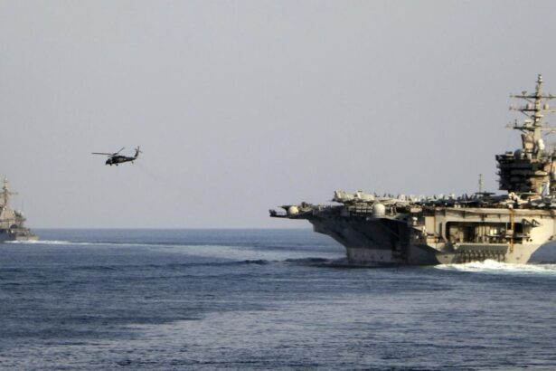 A massive U.S. aircraft carrier sailing across the sea while a helicopter closely follows it from the air