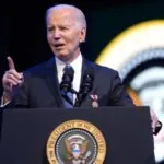 Biden wearing a handsome, elegant suit while speaking at a podium in front of many prominent people from the United States