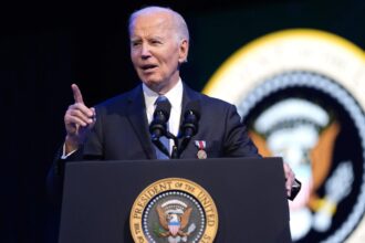 Biden wearing a handsome, elegant suit while speaking at a podium in front of many prominent people from the United States