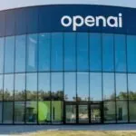 A panoramic view of OpenAI's headquarters featuring a modern glass façade with numerous windows and the company name displayed prominently in white on the building